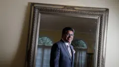 Una foto de Deepak Paradkar de 2013. Su reflejo es captado en un espejo con un marco plateado y ornamentado. Está mirando al espejo y sonriendo, lleva anteojos, un traje gris con una corbata rosa y un pañuelo de bolsillo a juego.