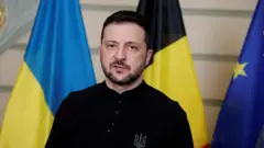 O presidente ucraniano Volodymyr Zelenskiy, vestido com uma camisa preta sem gola, retratado contra um fundo de bandeiras