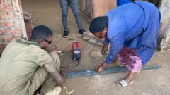 Françoise Umuhoza, wambaye itabuliya y'ubururu n'indorerwamo zijimye, agaragara ateranya ibyuma mu kazi ke akora ko gusudira.