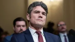 Joe Kent omuzdan yukarısı görülen fotoğrafta bir salonda oturuyor gerisinde birkaç resmi giyimli erkek flu. Hepsi karşılarına bakıyor. 