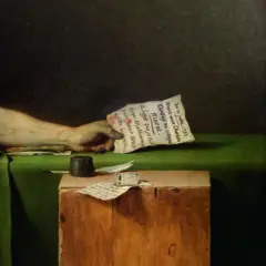 Un acercamiento a la mano del hombre muerto en la que sostiene una carta ensangrentada. En el cubo de madera que está al lado, se ve otra carta.