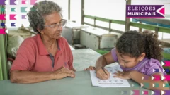 Seca na Amazônia: idosajogando roletapele escura e cabelo curtos sentada à mesa ensinando uma criança tambémjogando roletapela escura, que realiza atividade escrita