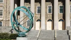 Um relógio de sol de metal no campus da Universidade Estadual da Pensilvânia, com o prédio Old Main ao fundo, um marco local em State College, Pensilvânia, EUA.