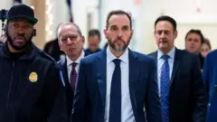 紺のスーツ姿のスミス氏が前をまっすぐ見据えて廊下を歩いている。隣に議会警官、周りに関係者や報道陣がいる