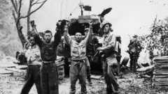 Três homens com as mãos levantadas enquanto um outro homem aponta uma arma para eles