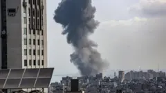 Coluna de fumaça ao lado de prédio e destroços