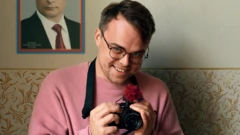 Мужчина держит в руках камеру и улыбается. На стене за ним портрет Владимира Путина.