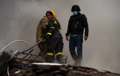 Yıkılan bir binada kurtarma ekipleri çalışıyor, bir kadın itfaiyeci hortum çekmeye çalışıyor. Yanında maskeli kişişler var.