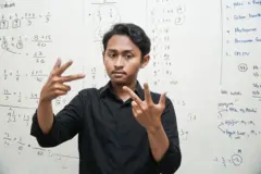 একটি সাদা বোর্ডের অঙ্কের নানা হিসাব লেখা, এর সামনে দাঁড়িয়ে আঙুল দিয়ে কিছু দেখাচ্ছেন কালো শার্ট পরা এক তরুণ- প্রতীকী ছবি।