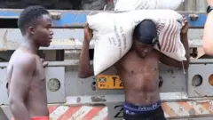 Abagabo batatu barimo gupakurura imifuka mu ikamyo. Babiri bari hasi batambaye mu gatuza, mu gihe imbere mu modoka inyuma hahagazemo undi mugabo