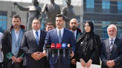 AKP'nin Üsküdar ilçe başkanı Sarıcaoğlu, 10 Ağustos Pazartesi günü Anadolu Adliyesi'nde basın açıklaması yapıyor. Etrafında diğer AKP'li üyeler var. Sarıcaoğlu kürsüye tutunuyor ve önünde ajanslara ait mikrofonlar var.