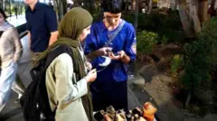 During a nighttime walk in northern Tehran, two young women are standing beside a small display of handmade pottery. One of them is entering information into a customer’s mobile phone. Pedestrians pass by, while the streetlights and surrounding trees illuminate the scene.