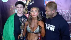 In front of a banner advertising a bodybuilding tournament, a tanned woman with well-defined muscles holds a trophy and is surrounded by bearded men and smiling young men.