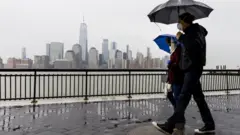 Duas pessoas usando máscaras caminham sob a chuva segurando guarda-chuvas com a cidade de nova york ao fundo, do outro lado do rio