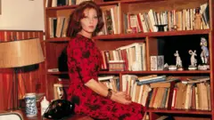 Clarice Lispector, com um vestido vermelho, em frente a uma estante de livros em seu apartamento no Leme em 1963