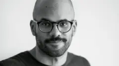 Kauê Lopes dos Santos, um homem careca, de barba curta e óculos, vestindo camiseta escura, em foto de rosto tirada com ângulo fechado