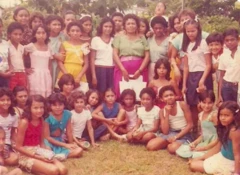 Dona Onete posa para foto cercada por crianças; ela veste uma camisa verde e uma saia rosa, e segura uma sacola.