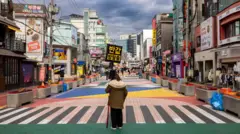 Uma rua de pedestres animada e colorida em Seul, repleta de faixas de pedestres vibrantes, lojas exclusivas e muita atividade. Uma mulher está no meio de uma faixa de pedestres, vestindo uma jaqueta de inverno e carregando a placa "Sala de Escape, metade do preço".