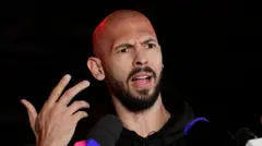 Andrew Tate wearing a black T-shirt, standing at a podium wit press microphones for im front