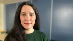 Stephanie Metzger vestindo blusa verde escura, com cabelo escuro longo, em frente a uma parede azul com linhas verticais. O fundo inclui à esquerda uma imagem desfocada do que parece ser uma mesa.