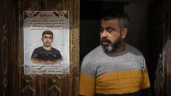 يقف والد جاد بجوار ملصق تذكاري لابنه، مُلصق على الباب الأمامي الخشبي المنحوت للعائلة في مخيم الفارعة للاجئين، في الضفة الغربية المحتلة