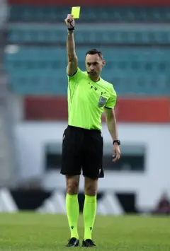 El árbitro Tom Owens, con camisa y medias verdes y pantaloneta negra, estira el brazo hacia arriba con una tarjeta amarilla en Estonia durante un partido el 4 de junio de 2026