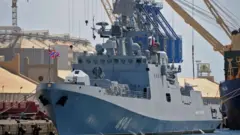 Російський військовий корабель сірого кольору, пришвартований у порту. На корпусі видно номер "494", на щоглі – російський прапор, а навколо розташовані портові крани та промислові споруди.