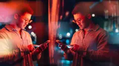 Um homem em pé sob luz vermelha, usando óculos e uma camisa, mexendo no celular em frente a um painel de vidro. Seu reflexo é mostrado do outro lado.