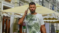 Загорелый мужчина в футболке разговаривает по телефону на улице Москвы. На заднем плане вывеска вареничной и зонтики над столами