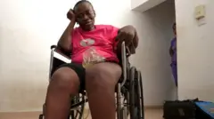 Muibat Onilude joko si ori kẹkẹ awọn alaabọ ara, o wọ asọ buba pupa ati sokoto penpe. O di iru adisẹyin, o si fi ọwọ osi le ẹrẹkẹ rẹ.