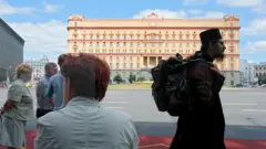 Pessoas passando pela calçada, incluindo um homem de roupa escura, em frente a um grande prédio amarelo no lado oposto da rua