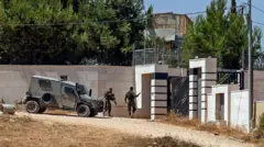 İki asker ve bir askeri araç lüks sayılabilecek bir evin kapısında duruyor. Askerlerden biri uzun namlulu silah taşıyor.