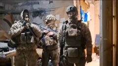 Троє військових у повному камуфляжному спорядженні та бронежилетах. Вони стоять у приміщенні з пошкодженими стінами, на яких видно папери та закріплені матеріали. Один із військових тримає автомат у руках, інші мають зброю на спорядженні. На задньому плані – стіл із речами та обладнанням. Освітлення штучне, загальний вигляд приміщення – імпровізований штаб або укриття.
