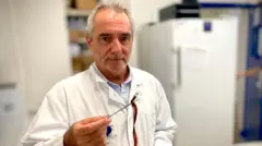 O professor Jean-Claude Alvarez aparece em seu laboratório de toxicologia, usando um jaleco branco. Ele tem cabelos grisalhos curtos e segura uma pinça com a qual prende alguns fios de cabelo.