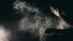 Dois braços abertos com as mãos espalmadas em fundo escuro, soltando partículas