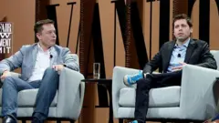 Elon Musk vestindo jeans azuis e uma jaqueta clara, sentado em uma poltrona, olhando em direção a Sam Altman, que está usando um terno escuro com tênis brancos, também sentado em uma poltrona. Entre eles há uma pequena mesa com um copo de água. Eles estão em um palco.
