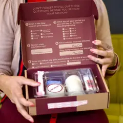 Uma mulher segura um kit de diagnóstico para uso em casa da NextGen. A caixa está aberta e, dentro, é possível ver as instruções de uso e os instrumentos que ajudam as usuárias a coletar suas amostras.