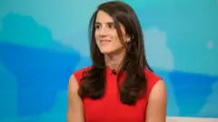 Kırmızı elbiseli siyah uzun saçlı bir kadın dik oturuyor ve bir kişiye bakıyor. Ortam bir televizyon stüdyosu izlenimi veriyor