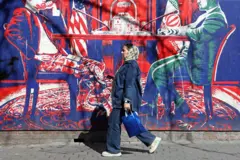 26 फरवरी, 2026 को तेहरान में पूर्व अमेरिकी दूतावास के बगल में स्थित ईरान-अमेरिका वार्ता को दर्शाने वाले अमेरिका विरोधी भित्तिचित्र के पास से एक ईरानी महिला गुजर रही है.