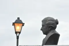 Busto de Cora Coralina com luminária antiga ao fundo