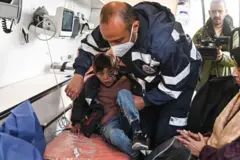 صبي فلسطيني يجلس فيما يبدو أنه سيارة إسعاف، يمسكه مسعف، وبجانبه سيده، وخلفهم صحفي يصور المشهد بكاميرا فيديو سوداء