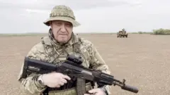 Yevgeny Prigozhin aparece no deserto, vestido com uniforme militar, de chapéu, segurando uma arma