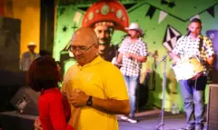 Casal dançando forró com uma dupla de músicos desfocada ao fundo, em frente a um mural com a imagem de um homem usando chapéu de cangaceiro
