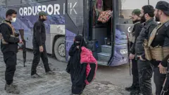 Silahlı kişilerin gardiyanlığında bir genç kız otobüsten iniyor. Tamamen çarşaflı bu kızın elinde pembe bir mont var.