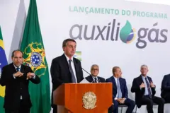 Bolsonaro discursa em um púlpito da presidência da República, com integrantes do governo sentados atrás. Atrás de Bolsonaro, há também um banner com a mensagem: 'Lançamento do programa Auxílio Gás".