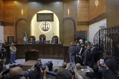 قاعة محكمة القصر العدلي في دمشق ممتلئة، ويظهر في مقدمة القاعة منصة يجلس عليها ثلاثة قضاة، وفوقهم ميزان العدالة وآية قرآنية. وعلى يمين القاعة قفص اتهام يقف داخله متهم واحد، إلى جانب حضور إعلامي وجمهور يتابع مجريات الجلسة.