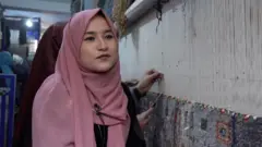 صالحه حسنی، ۱۹ ساله لباس سیاه و چادر مرجانی رنگ، در کنارش یک دستگاه قالین بافی است که در عقب او چند دختر در حال بافتن قالین هستند.