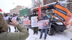 На заснеженной улице стоит группа людей с плакатами и флагами возле припаркованных автомобилей. На одном из авто закреплен большой муляж ракеты с надписью «На Вашингтон», а рядом виден черно-оранжевый флаг. Люди держат различные лозунги, направленные против США и в поддержку российской власти. На переднем плане женщина в зимней куртке фотографирует эту сцену на телефон.