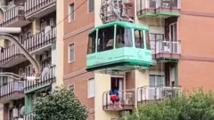 街中を通るロープウェーの明るい緑色のゴンドラから、長髪の人物がロープで降ろされている写真