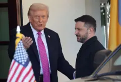 Trump cumprimenta Zelensky na entrada da Casa Branca e diz algo em tom leve. Zelensky sorri. No primeiro plano, aparece a bandeira dos EUA na frente de um carro.